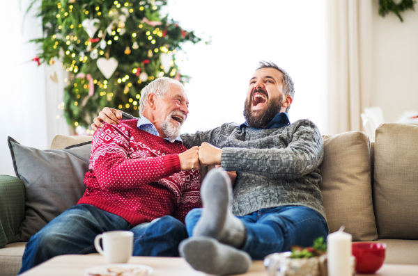 A senior father and adult son with feet on a coffee table sitting on a sofa at home at Christmas time, laughing.