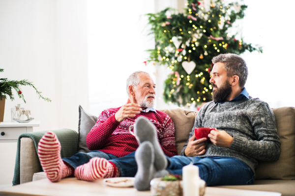 A senior father and adult son with feet on a coffee table sitting on a sofa at home at Christmas time, talking.