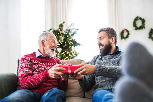 A senior father and adult son sitting on a sofa at home at Christmas time, giving presents to each other.