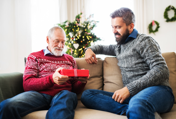 A senior father and adult son sitting on a sofa at home at Christmas time, giving presents to each other.