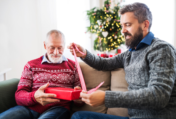 A senior father and adult son sitting on a sofa at home at Christmas time, wrapping up a present.