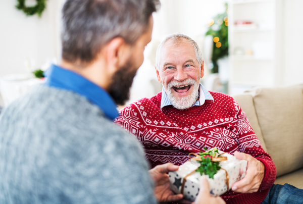 A senior father and adult son sitting on a sofa at home at Christmas time, giving presents to each other.