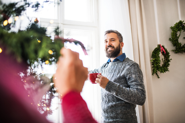 A hipster man standing at a window with a red cup at home at Christmas time, talking to an unrecognizable person.