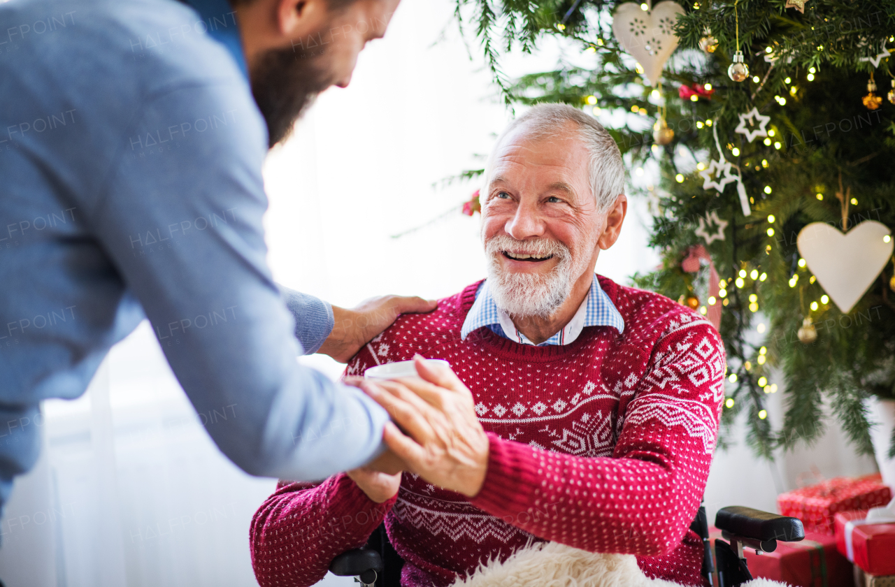 An unrecognizable hipster man giving a drink in a cup to his senior father in wheelchair at Christmas time.