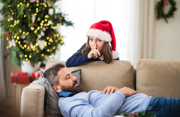 A small girl with Santa hat doesn't want to wake up her sleeping father at Christmas time.