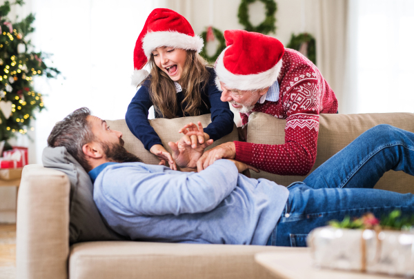 A small girl and her grandfather with Santa hat waking up her father sleeping on a sofa at Christmas time.