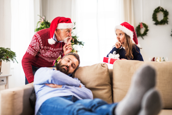 A small girl with Santa hat and present and her grandfather don't want to wake up her sleeping father at Christmas time.