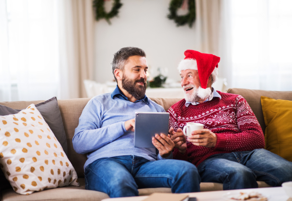 A senior father and adult son with tablet sitting on a sofa at home at Christmas time, talking.