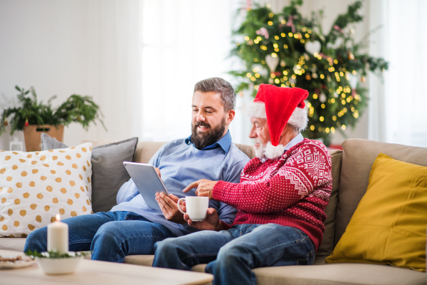 A senior father and adult son with tablet sitting on a sofa at home at Christmas time, talking.