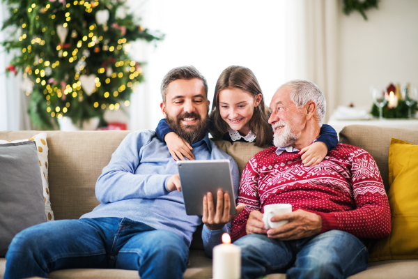 A happy small girl and her father and grandfather sitting on a sofa at Christmas time, using tablet.