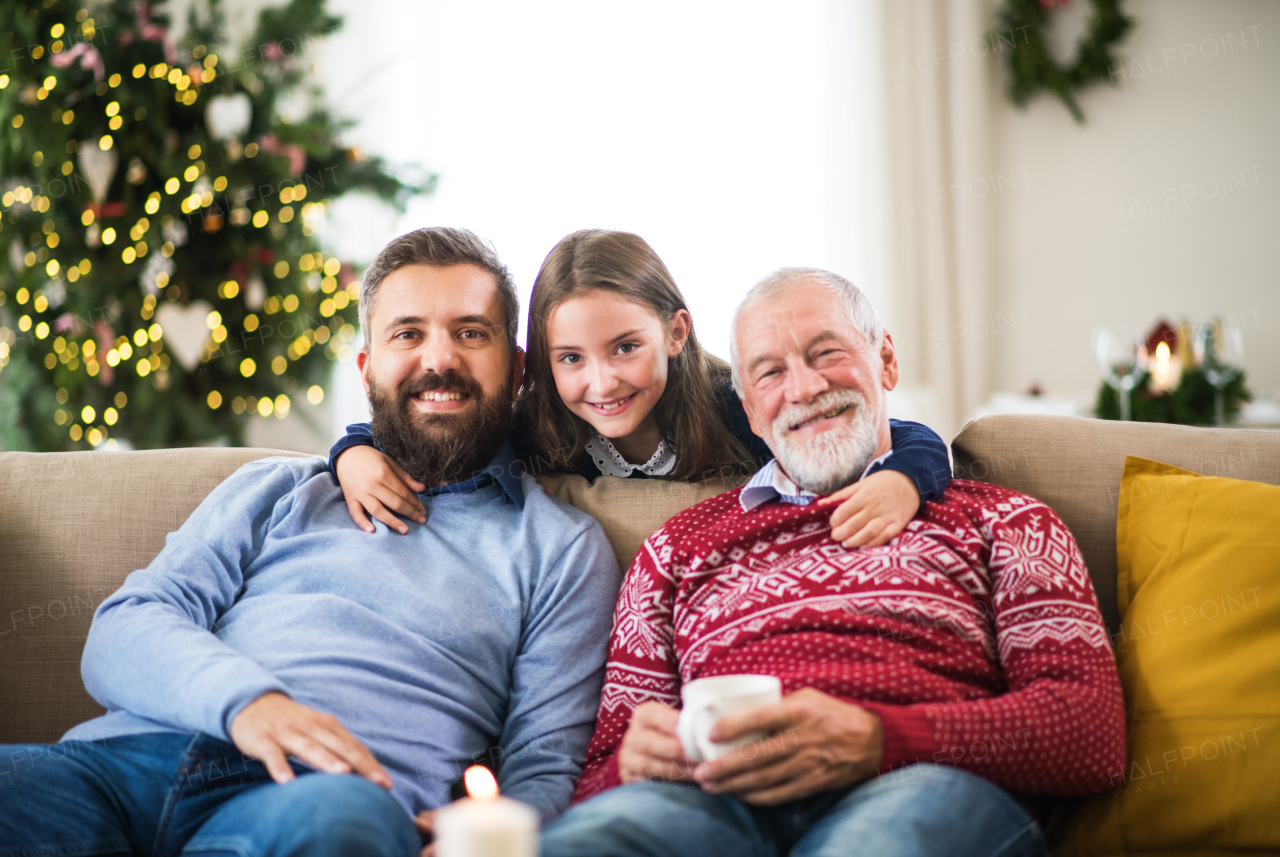 A happy small girl and her father and grandfather sitting on a sofa at Christmas time, posing for the photograph.