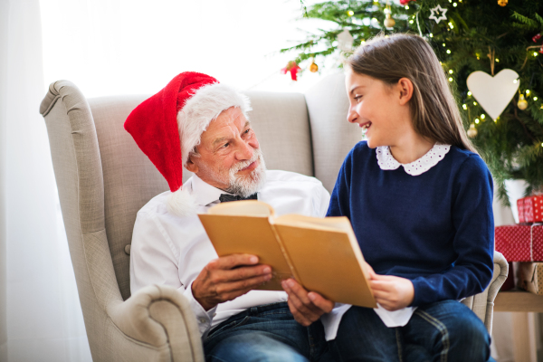 A small girl and her grandfather with Santa hat sitting on armchair and reading a book at Christmas time.