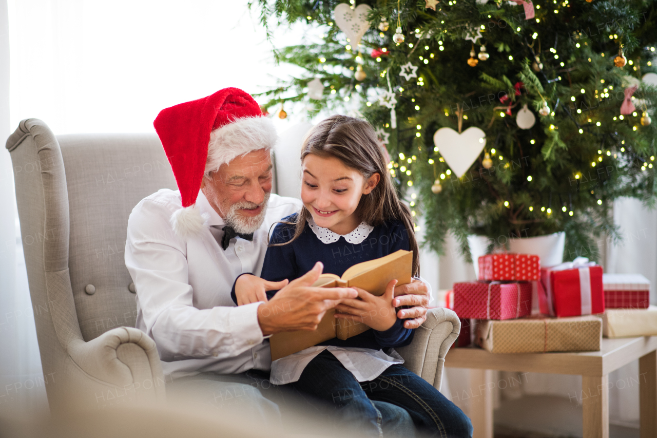 A small girl and her grandfather with Santa hat sitting on armchair and reading a book at Christmas time.