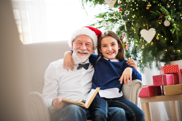 A small girl and her grandfather with Santa hat sitting on armchair and reading a book at Christmas time.