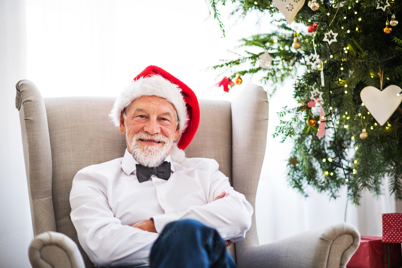 A senior man with a Santa hat sitting on an armchair at home at Christmas time.