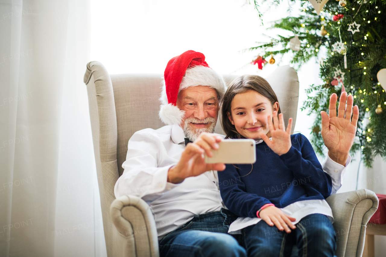 A happy small girl and her grandfather with Santa hat taking selfie with smartphone at Christmas time, waving.