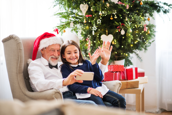 A happy small girl and her grandfather with Santa hat taking selfie with smartphone at Christmas time, waving.