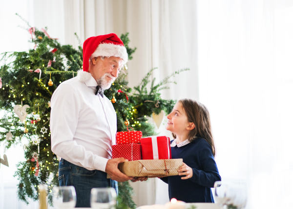 A senior man with a Santa hat giving presents to a happy small girl at Christmas time.