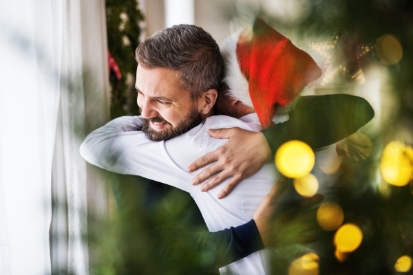 A senior father with a Santa hat and adult son standing by the window at home at Christmas time, hugging.