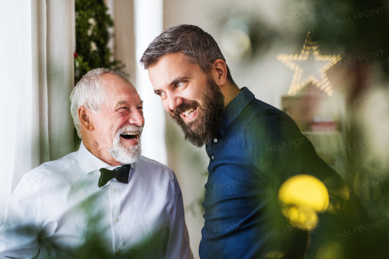 A senior father with a bowtie and adult son standing by the window at home at Christmas time, looking out and talking.