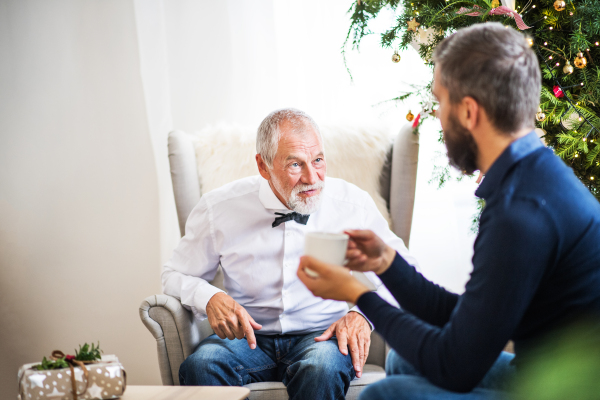 A senior father and adult son with a cup of coffee sitting on a sofa at Christmas time, talking.