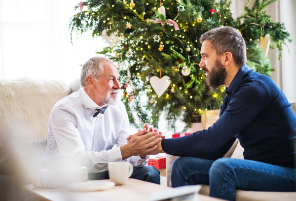 A senior father and adult son sitting on a sofa at Christmas time, looking at each other, holding hands and talking.