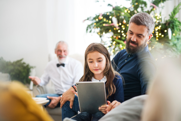 A small girl with tablet and her father and grandfather sitting on a sofa at Christmas time.