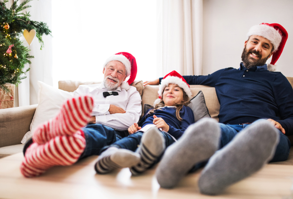 A small girl with father and senior grandfather sitting on a sofa at Christmas time, feet on a table and Santa hats on their head.