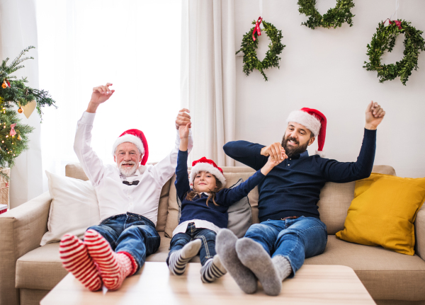 A small girl with father and senior grandfather sitting on a sofa at Christmas time, feet on a table and Santa hats on their head.