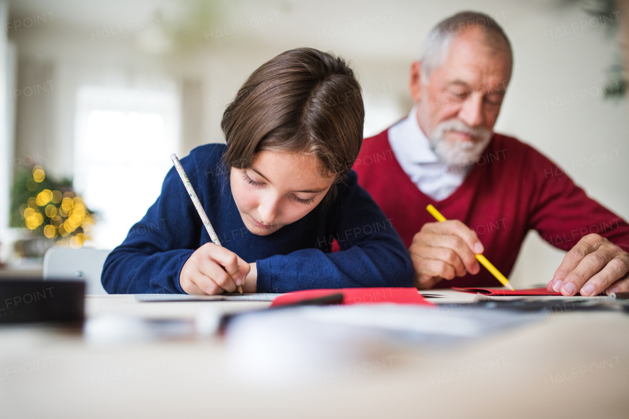 A small girl and her grandfather sitting at a table, writing Christmas cards together.