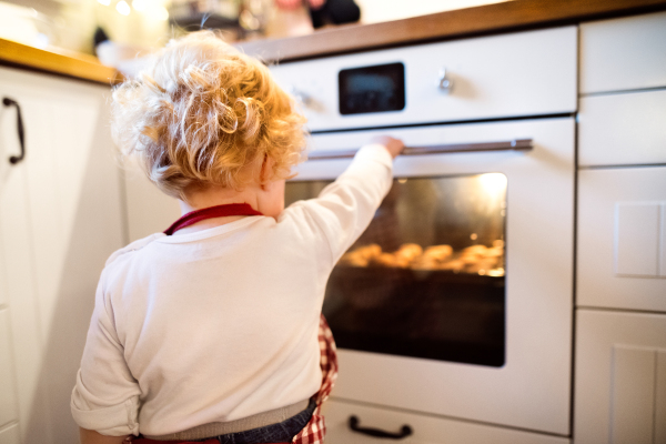 Young family making cookies at home. Toddler boy looking at cookies in the oven. Rear view.