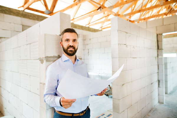 Young hipster architect or home owner at the construction site looking at blueprints, controlling issue.