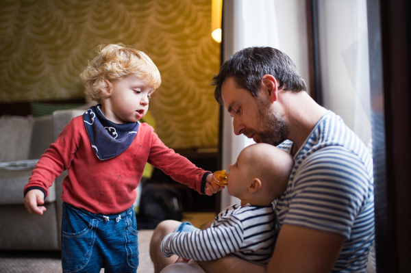 Father with a toddler boy and a baby in a hotel, summer day. Young man with children sitting on the floor inside the room.