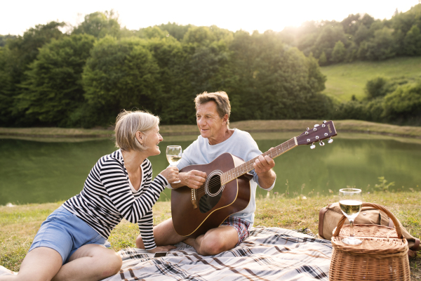 Beautiful senior couple at the lake having a picnic, sitting on blanket, drinking wine. Man playing guitar.