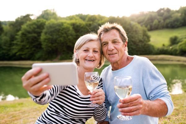 Beautiful senior couple at the lake having a picnic, drinking wine, holding smart phone, taking selfie.