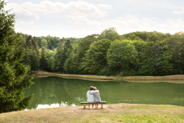 Beautiful senior couple on a walk at the lake sitting on wooden bench hugging. Rear view.