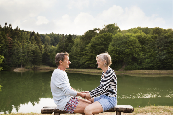 Beautiful senior couple on a walk at the lake sitting on wooden bench, holding hands.