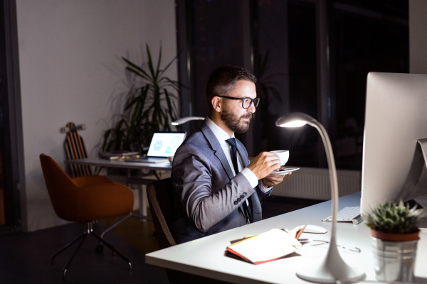 Handsome hipster businessman in his office late at night, sitting at the desk, computer in front of him, drinking coffee.