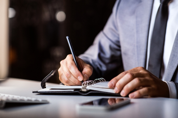 Unrecognizable businessman in his office late at night, sitting at the desk, writing something into his personal organizer, smart phone laid next to him.