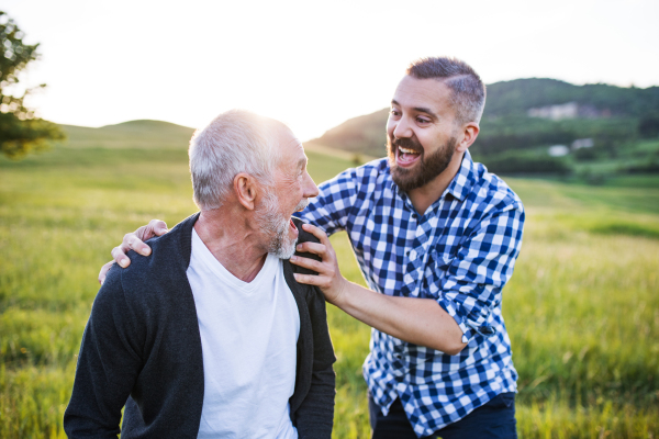 An adult hipster son with his senior father on a walk in nature at sunset, having fun.