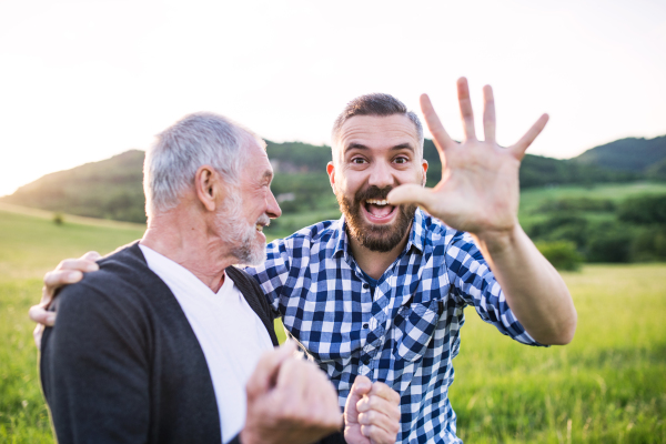 An adult hipster son with senior father on a walk in nature at sunset, having fun.