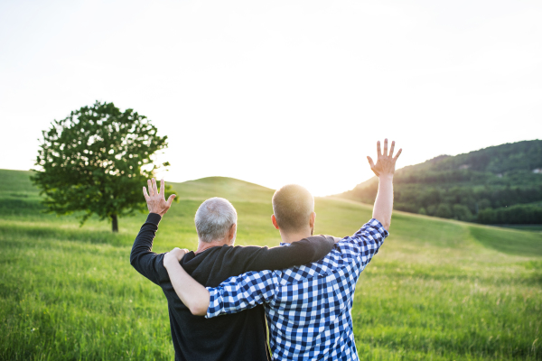 An adult hipster son with his senior father on a walk in nature at sunset, arms around each other. Rear view.