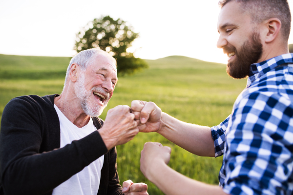 An adult hipster son with his senior father making fist bump in nature at sunset. Close up.