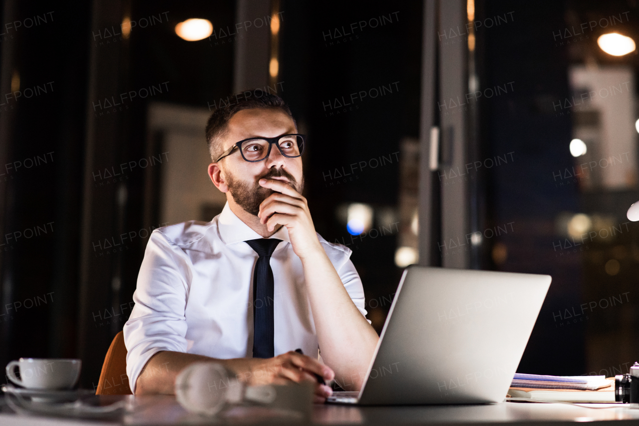 Thoughtful hipster businessman in his office working late at night, sitting at the desk, laptop in front of him.