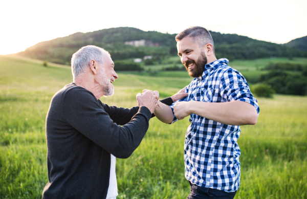 An adult hipster son with his senior father making fist bump in nature at sunset. Close up.