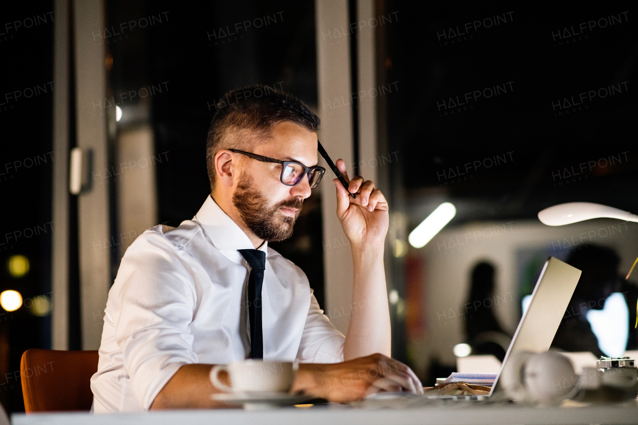 Thoughtful hipster businessman in his office working late at night, sitting at the desk, laptop in front of him.