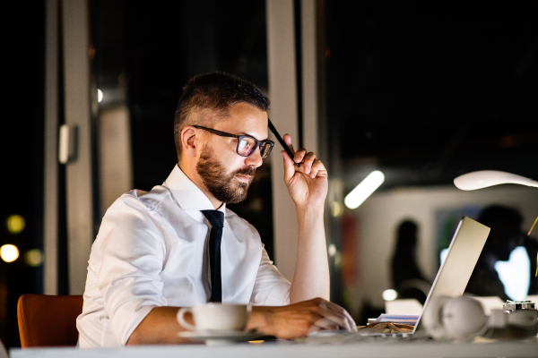 Thoughtful hipster businessman in his office working late at night, sitting at the desk, laptop in front of him.