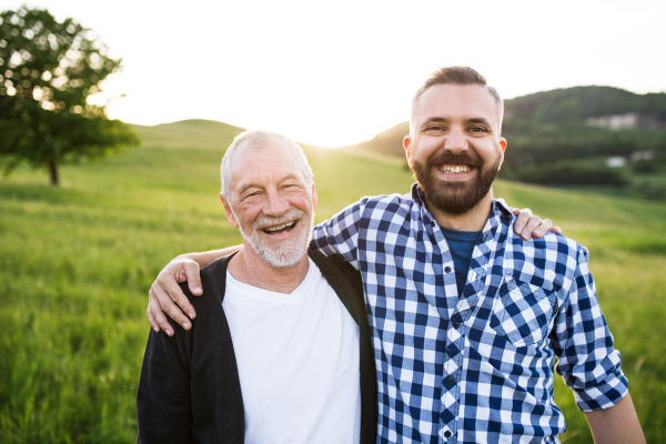 A portrait of a laughing adult hipster son with senior father in nature at sunset, arms around each other.