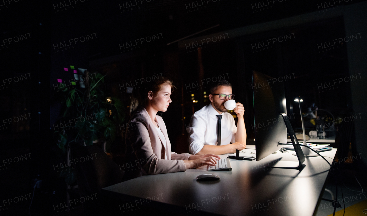 Two young businesspeople in the office at night working late, discussing a project, drinking coffee.