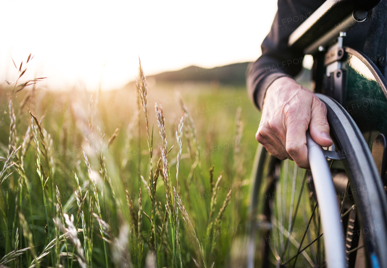 A close-up of senior man's hand on a wheelchair in nature at sunset. Copy space.